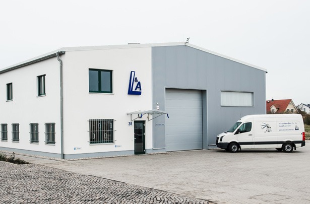 Außenansicht eines modernen Firmengebäudes mit dem Logo "L&A". Vor dem Gebäude ist ein Firmenfahrzeug geparkt, die den mobilen Service des Unternehmens für Laserschweißen und ähnliche Dienstleistungen repräsentieren. Der große gepflasterte Hof bietet viel Platz für Anlieferungen oder Kundenfahrzeuge.