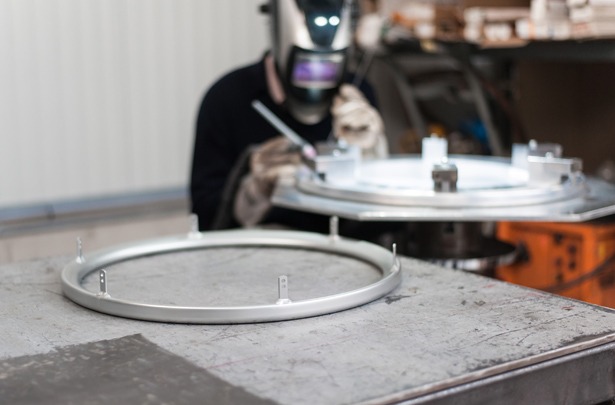Ein Schweißer mit Schweißhelm arbeitet präzise an einem runden Metallwerkstück Schweißgeräts. Das Werkstück ist auf einem drehbaren Tisch befestigt, um exakte Schweißnähte zu ermöglichen. Im Vordergrund liegt ein fertig geschweißtes Bauteil.