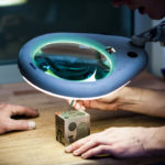 Qualitätskontrolle der Lasergravur unter einer großen Lupe durch zwei Mitarbeiter.