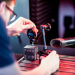 Ein Techniker justiert ein Messgerät an einem Metallblock, um die Genauigkeit bei der Lasergravur sicherzustellen. Im Hintergrund ist eine Absauganlage zu sehen, die den Arbeitsplatz sauber hält.