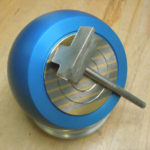 Blaue, hart eloxierte Magnetspannkugel mit Ø 140–160 mm bringt ein rechteckiges Metallbauteil in Position: Das Werkstück sitzt mittig auf der magnetischen Spannfläche mit feinen, konzentrischen Rillen; ein T‑Griff steht schräg im eingelegten Schlüssel­schlitz. Die Kugel ruht auf einer silberfarbenen Abschlussplatte