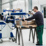 Ein mobiles Laserschweißgerät in einer Werkstattumgebung. Ein Techniker arbeitet konzentriert mit einem Mikroskop, um präzise Schweißnähte an einem Werkstück zu erstellen. Das robuste Gerät ist auf Rollen montiert und für den flexiblen Einsatz geeignet.