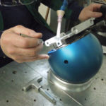 Blaue Magnetspannkugel im Einsatz: Die Kugel fixiert ein Werkstück (zylindrisches Metallteil) sicher auf einer Laserbearbeitungs- oder Polierplatte. Der T-Griff ist sichtbar herausgezogen, um die magnetische Haltekraft auszuschalten, während Metallspäne und reflektierende Oberflächen der Anlage im Hintergrund auf den industriellen Einsatz hinweisen.