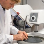 Mitarbeiter im weißen Laborkittel poliert eine Werkstoffprobe manuell auf einer Schleif- und Poliermaschine.