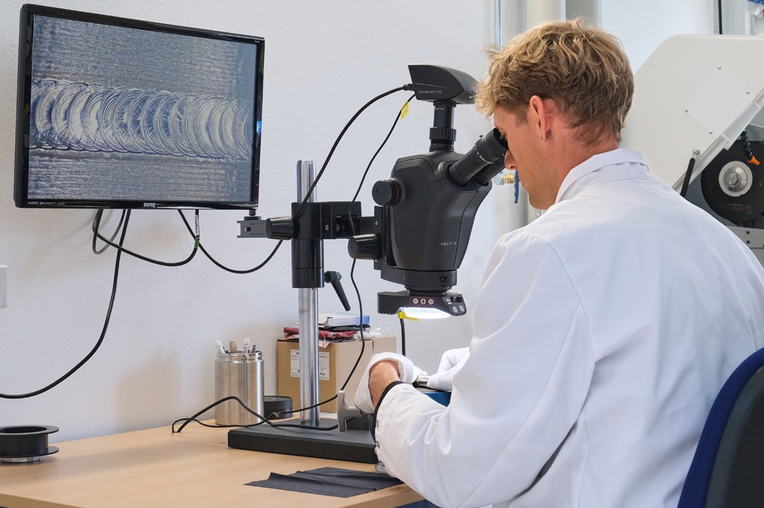 Mitarbeiter im weißen Laborkittel untersucht unter einem Stereomikroskop die Oberfläche eines Werkstoffs; das vergrößerte Bild wird auf einem Monitor angezeigt – Teil der zerstörenden Werkstoffprüfung (Makroschliff).