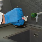 Hand mit blauen Laborschutzhandschuhen hält eine Werkstoffprobe mit aufgetragener Ätzlösung unter der Laborabzugshaube.