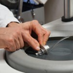 Hände eines Mitarbeiters führen eine metallische Probe über eine rotierende Polierscheibe mit Wasserkühlung.